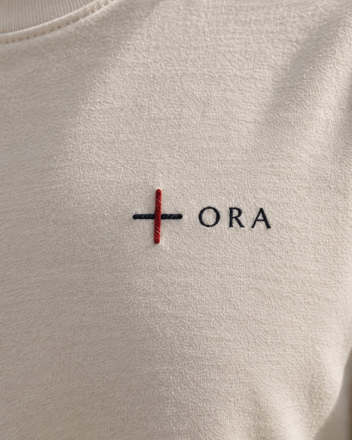 Klein borduursel op de borst van een T-shirt: het kruis van Ink en scharlaken en het woord ORA